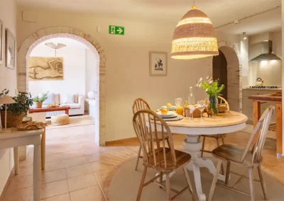 Palmera family apartment's kitchen- dining area has the table laid for breakfast and fresh flowers picked in the grounds. You can see the bright and spacoius lounge through the brick arch